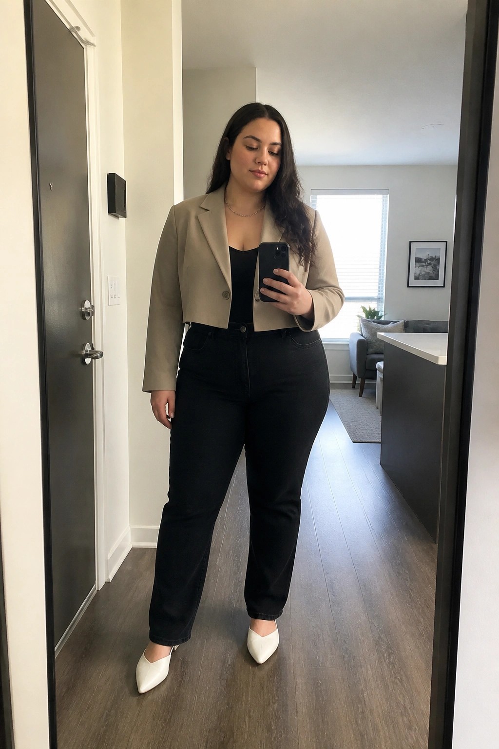 Woman in beige cropped blazer and black jeans taking mirror selfie in apartment