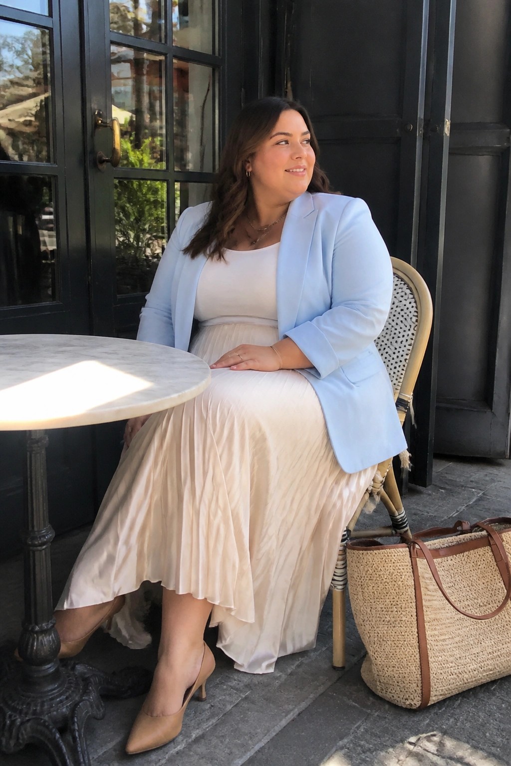 Woman in light blue blazer and beige pleated skirt beside straw tote at marble table