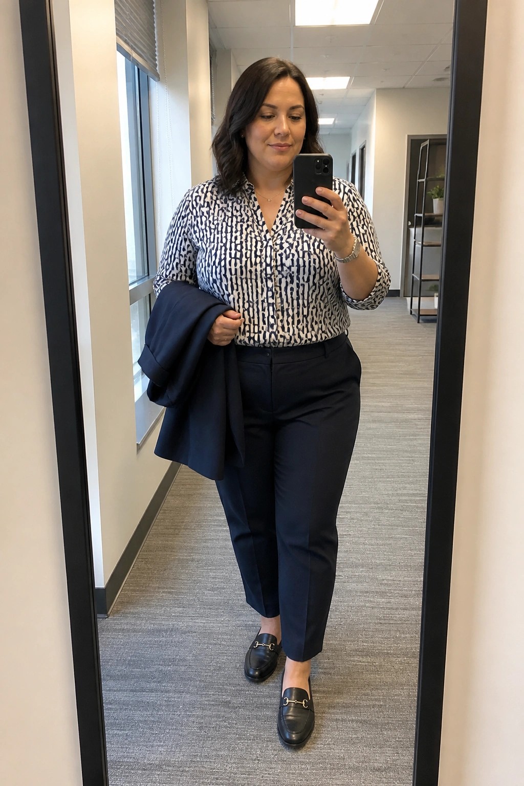Woman in plus size navy trousers and black white patterned button down blouse holding a navy blazer over one arm with black loafers and minimal jewelry in an office hallway.