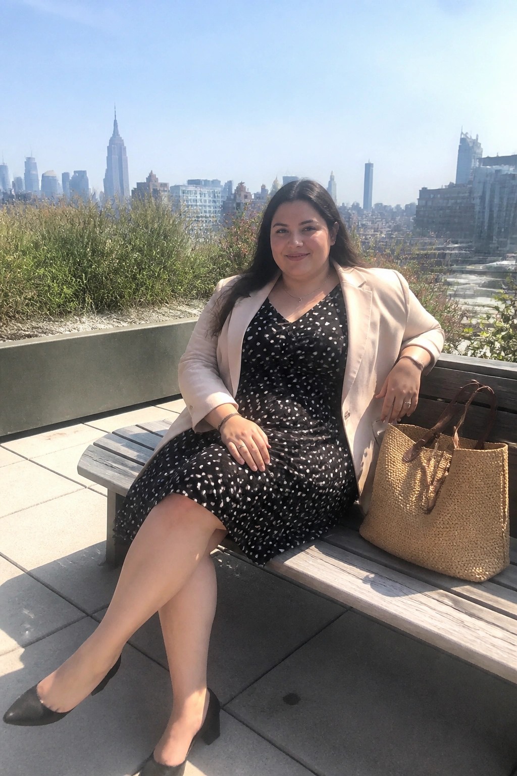 Woman in polka-dot dress and beige blazer on bench with straw tote, NYC skyline beyond.