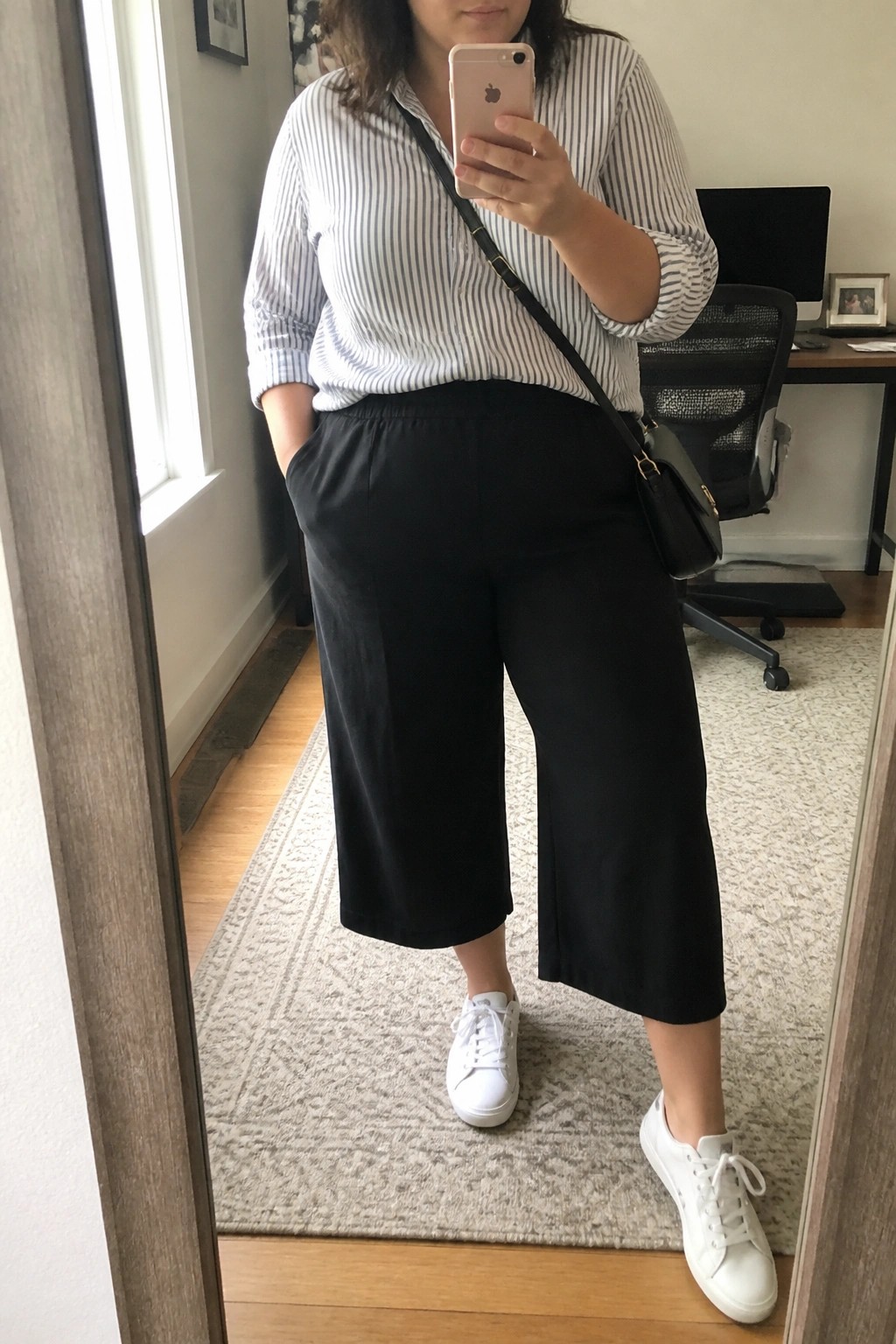 Plus size woman wearing a white and blue striped button down shirt with black wide leg cropped pants, white sneakers, and a black crossbody bag while holding a phone.