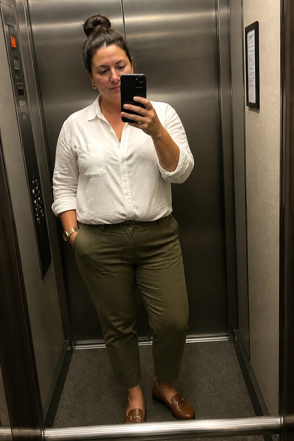Plus size woman wearing a white long sleeve button down shirt tucked into olive green straight leg pants with brown loafers and a gold watch while taking a mirror selfie in an elevator.