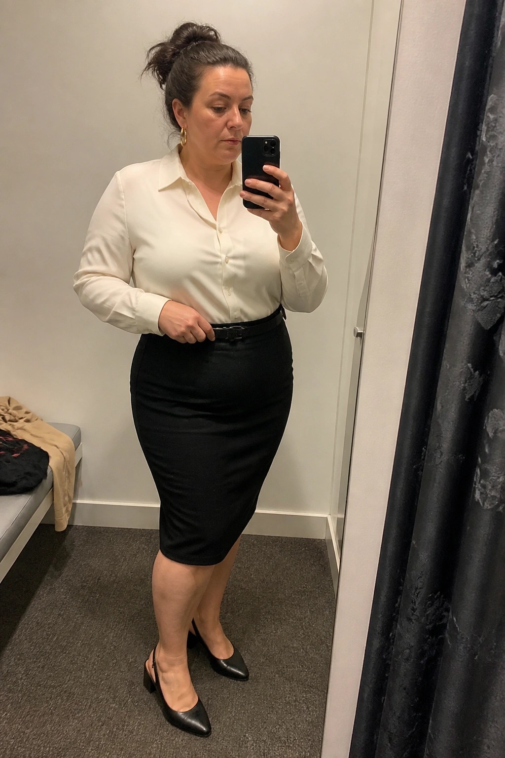 Woman in white shirt and black pencil skirt taking mirror selfie in fitting room
