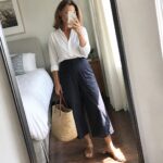 Woman in white shirt and navy culottes taking mirror selfie with straw bag.