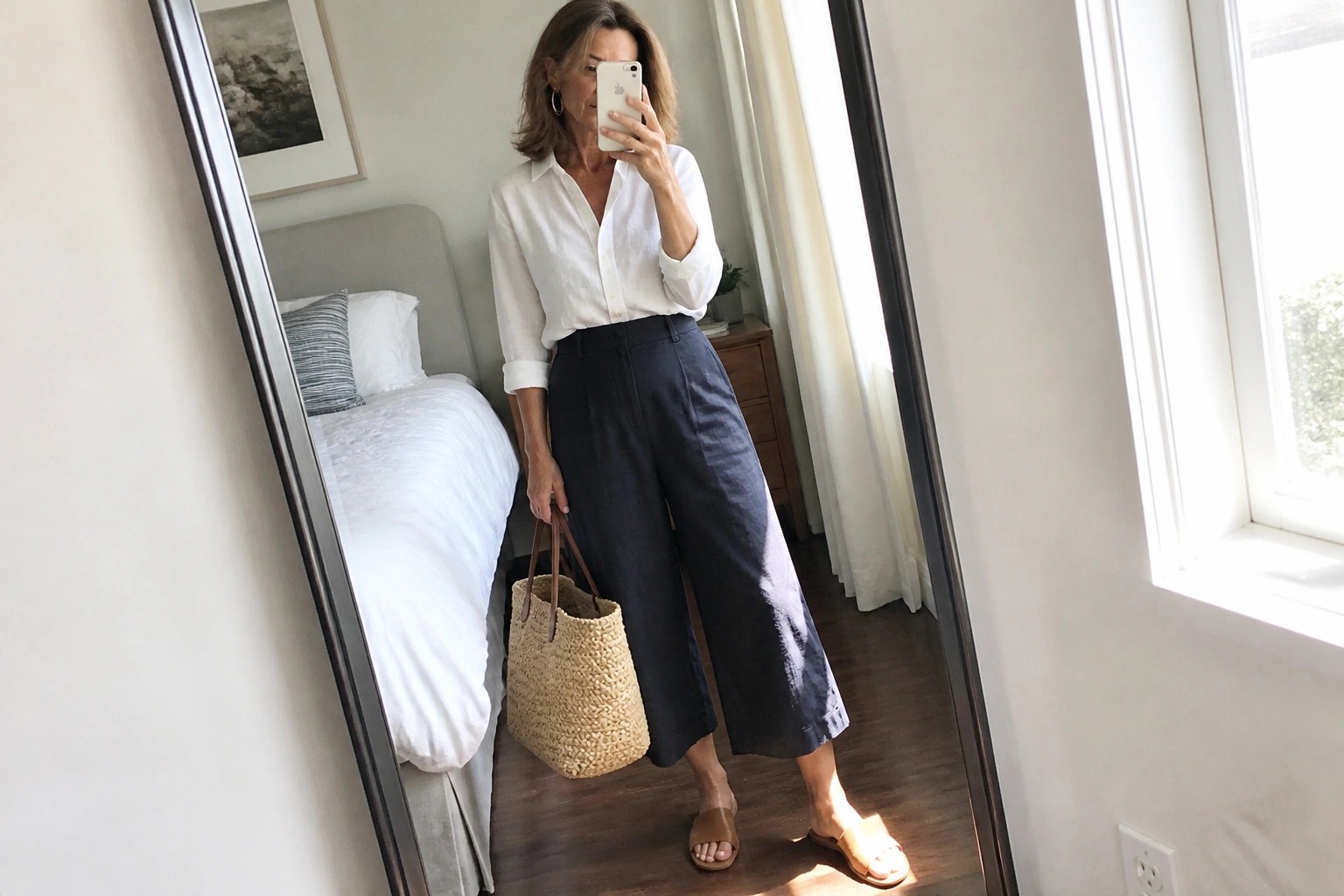 Woman in white shirt and navy culottes taking mirror selfie with straw bag.