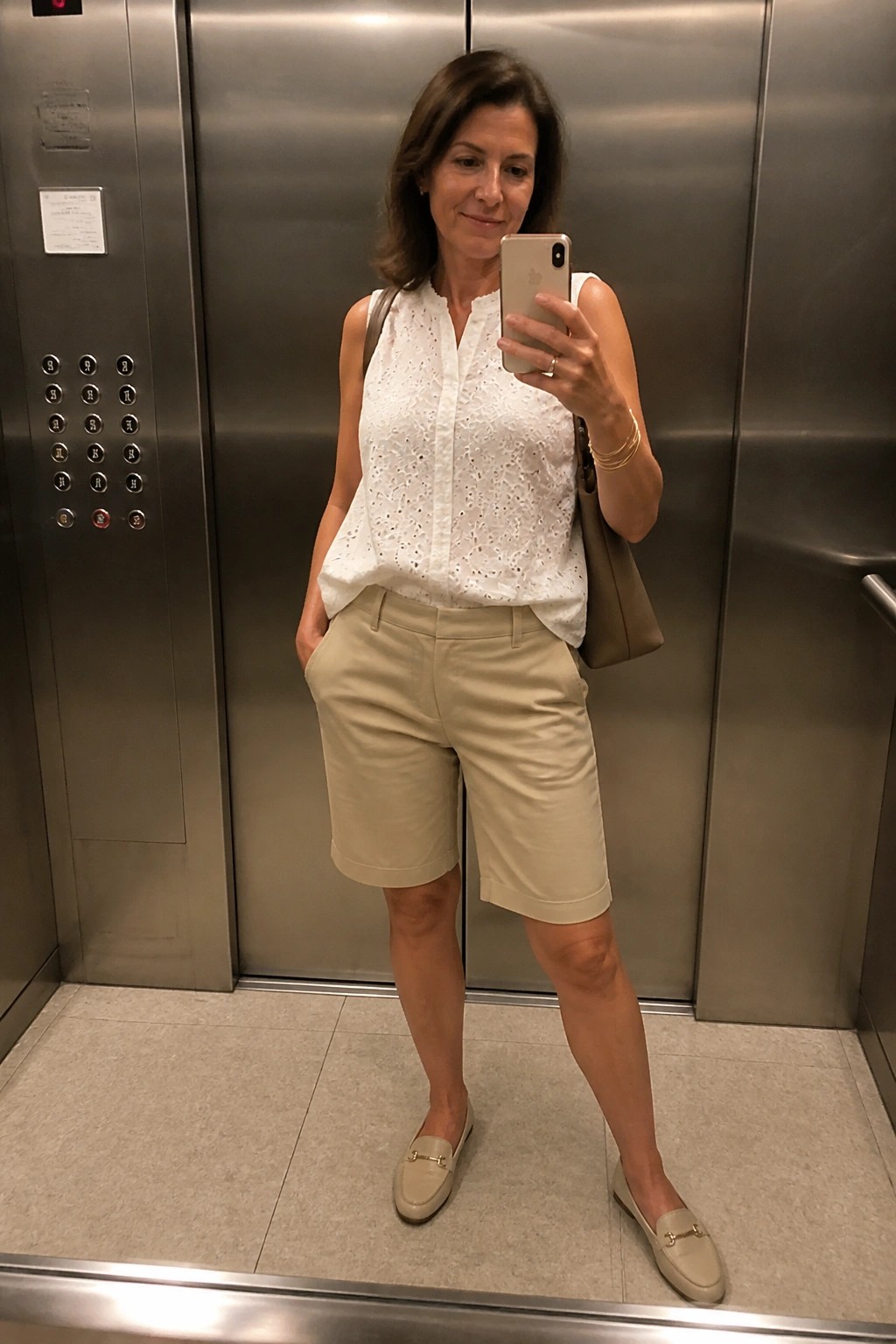 Woman in white eyelet top and beige shorts taking selfie in elevator