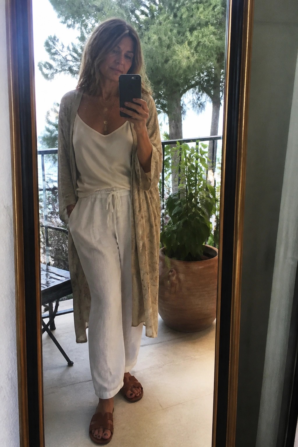 Woman in white linen outfit and beige cardigan taking balcony mirror selfie with phone