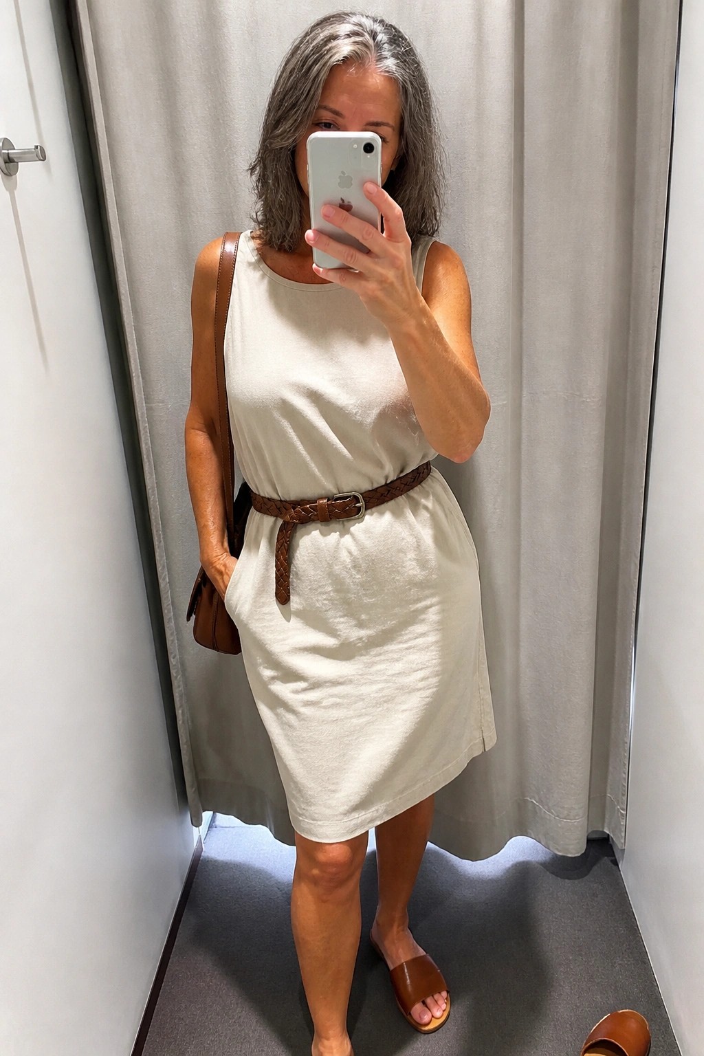 A woman over 50 wearing a cream sleeveless shift dress cinched with a brown braided leather belt, paired with brown leather slides and a shoulder bag, taking a mirror selfie.