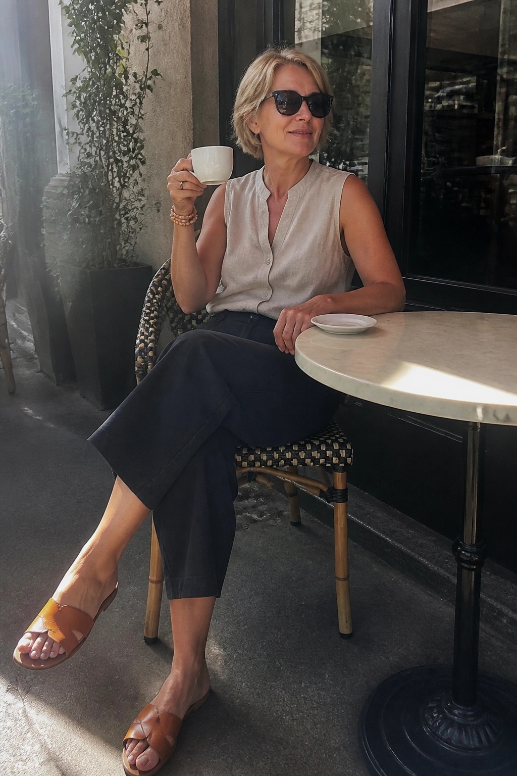 Woman in beige sleeveless top holds white cup at sunny outdoor cafe table