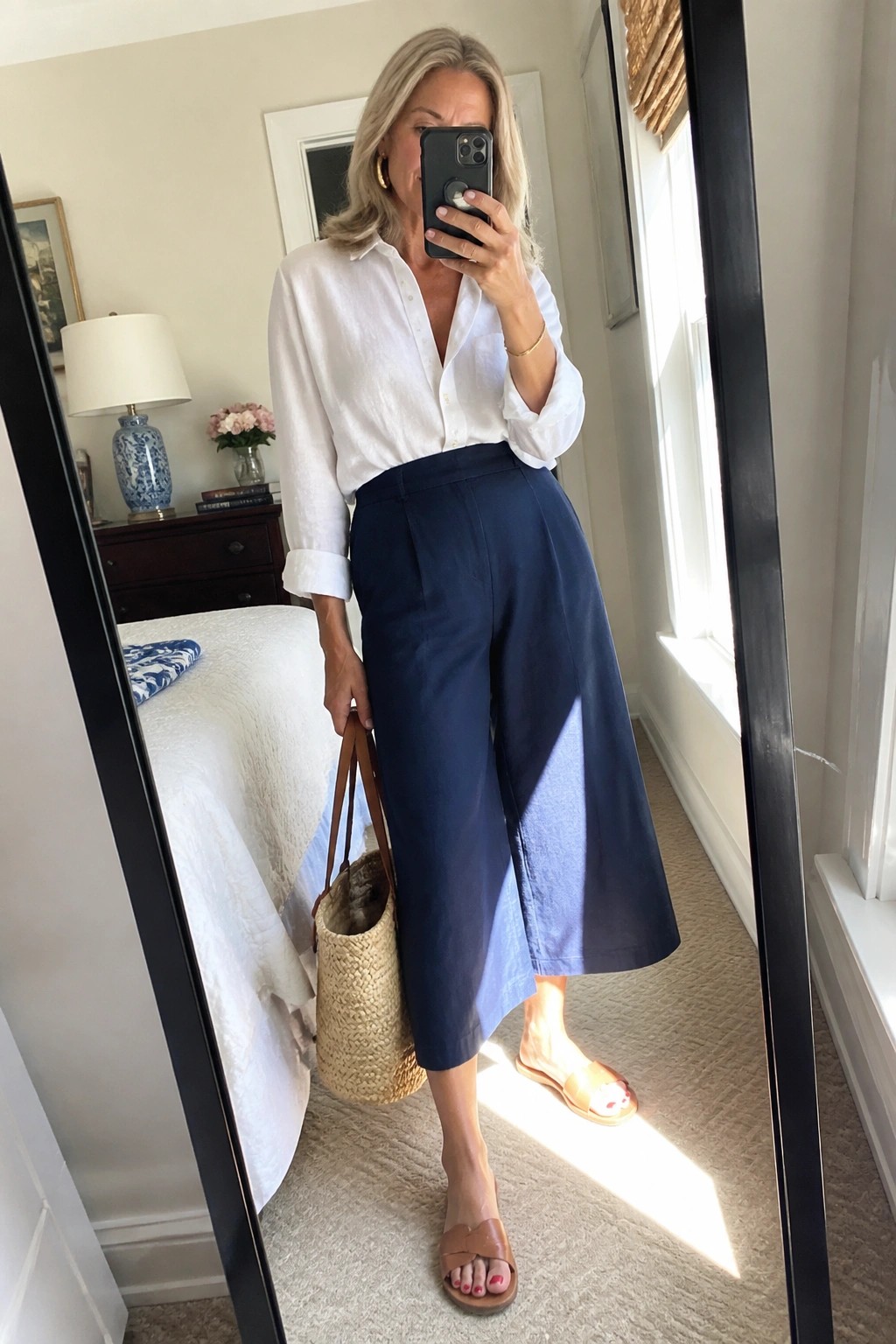 Woman in white shirt and navy culottes taking mirror selfie with straw bag.
