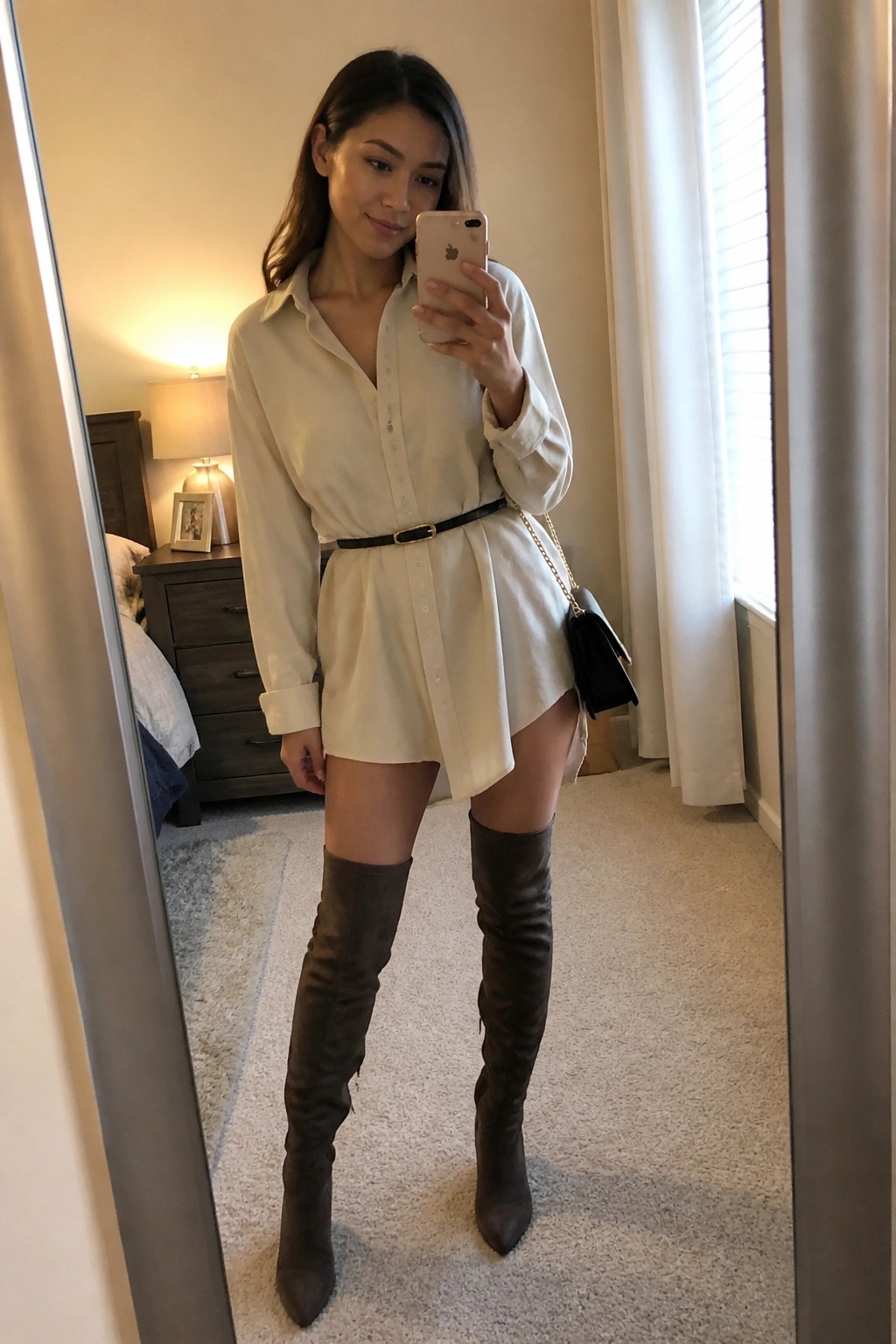 Woman in white shirt dress and brown over-the-knee boots taking mirror selfie in bedroom