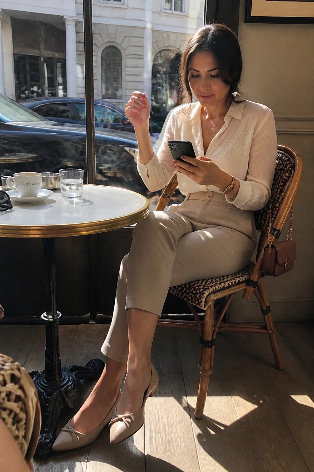 Woman wearing a cream button down blouse tucked into taupe cropped trousers with beige pointed toe flats and delicate jewelry while seated at a cafe table.