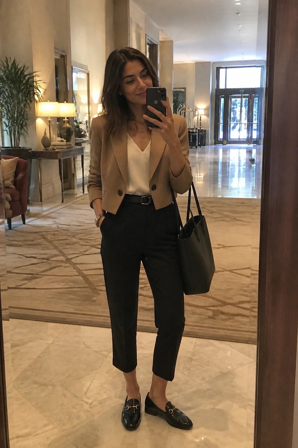 Woman in tan blazer, white top, and black pants taking mirror selfie in lobby