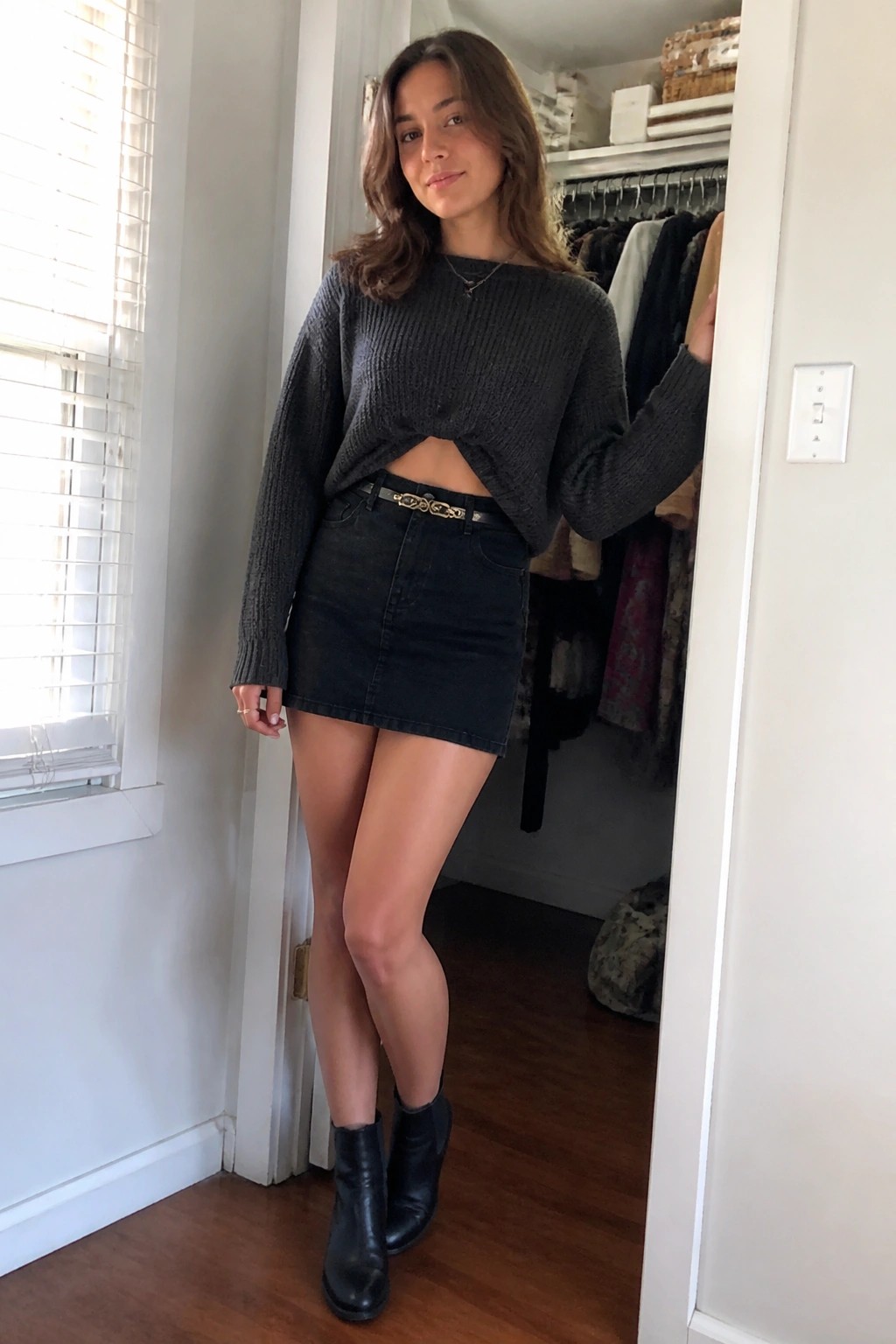 Woman in cropped gray sweater, black denim mini skirt, and ankle boots in doorway