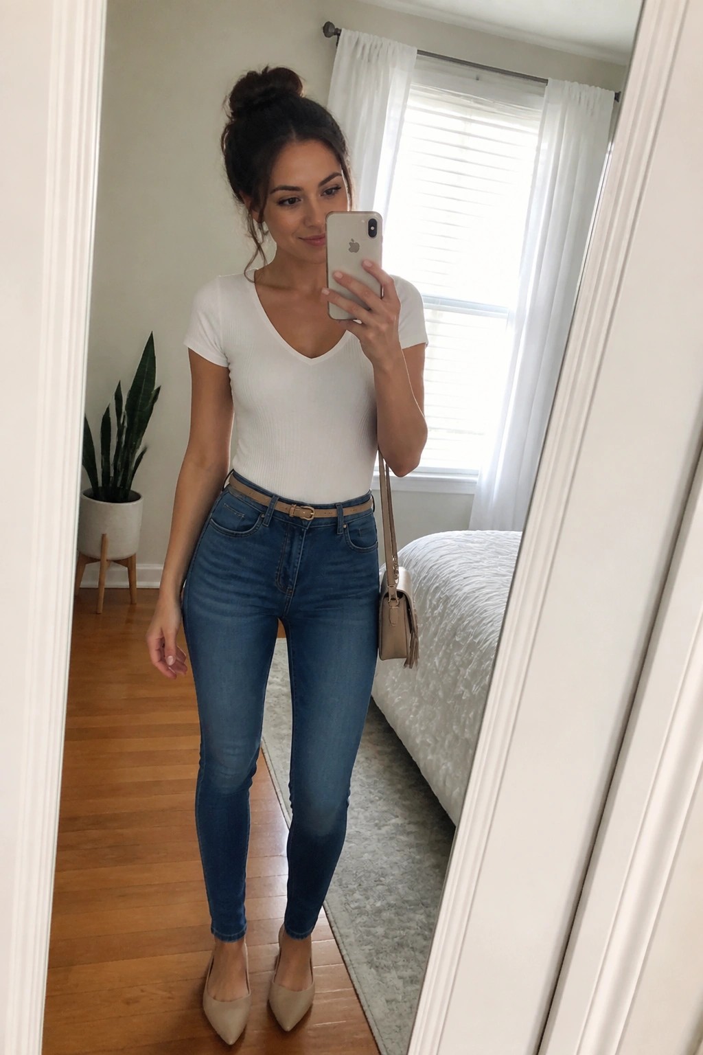 Woman in white ribbed V-neck top tucked into high-waisted blue skinny jeans with beige belt, beige pointed flats, and small crossbody bag, taking a mirror selfie.