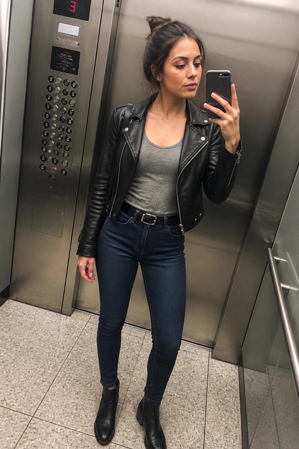 Woman in black leather jacket and jeans taking mirror selfie in elevator.