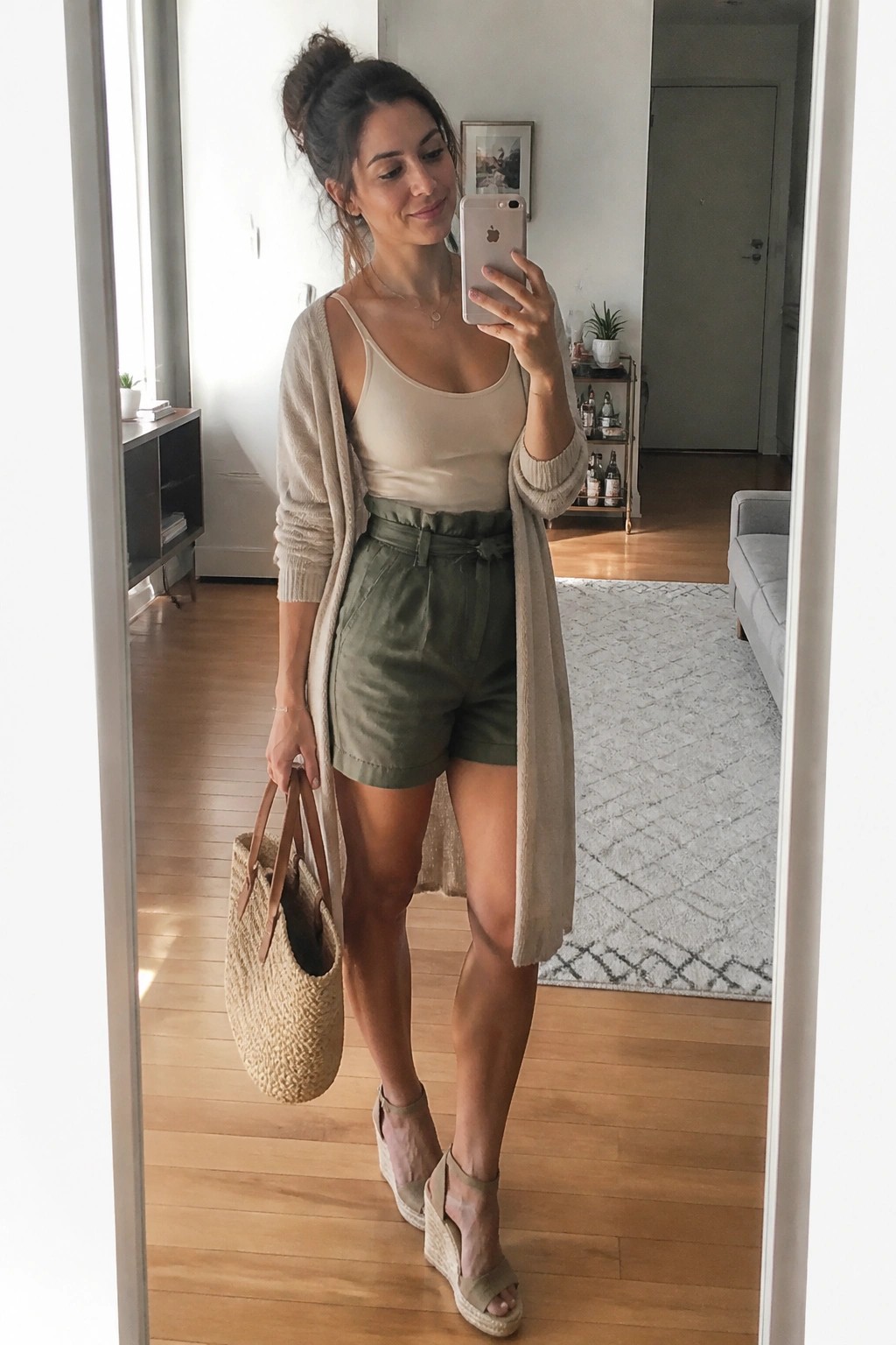 Woman in beige cardigan and olive shorts holding woven bag for mirror selfie