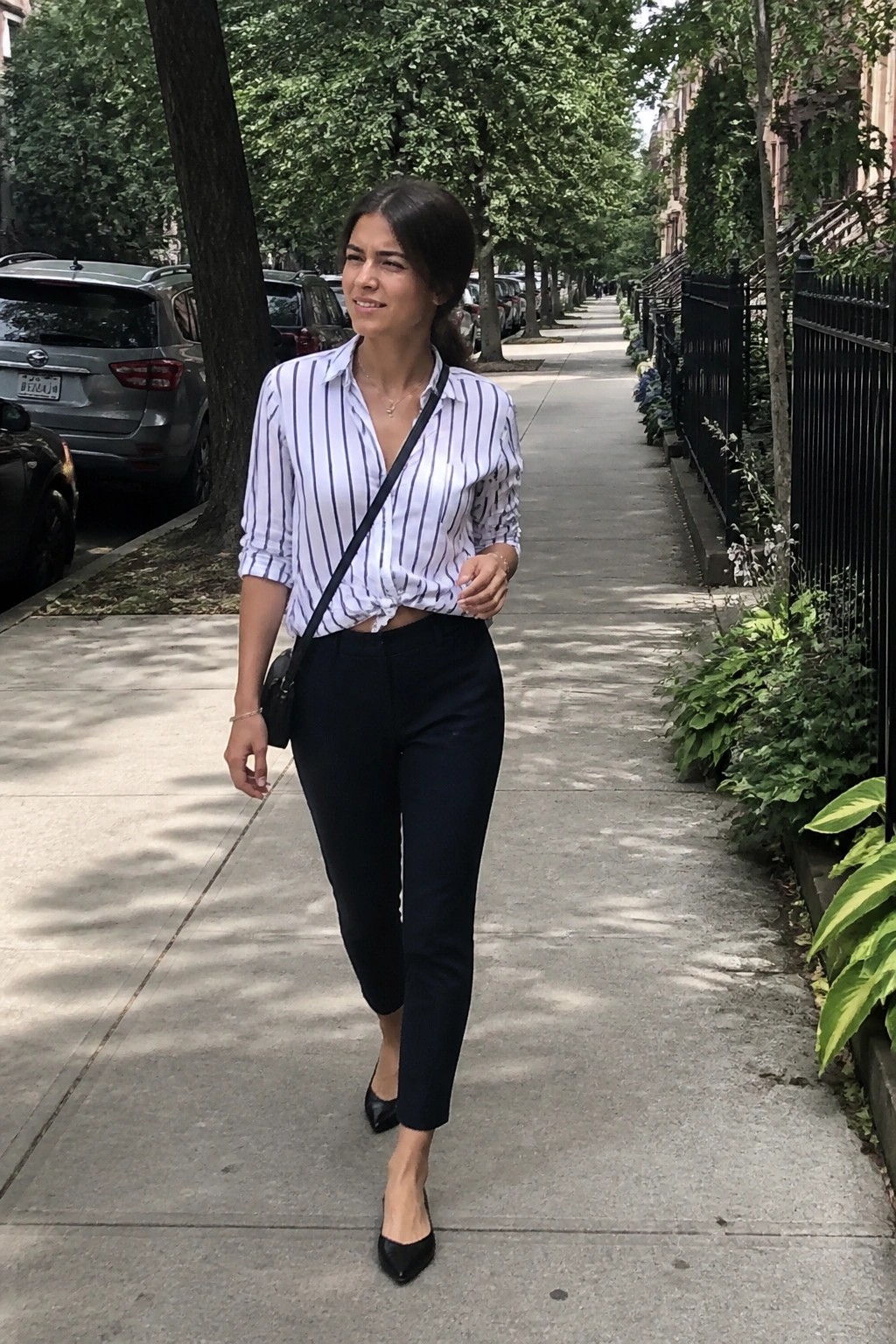 Woman wearing light blue and white striped button down shirt tied at waist with black cropped pants and black pointed flats plus thin crossbody bag.
