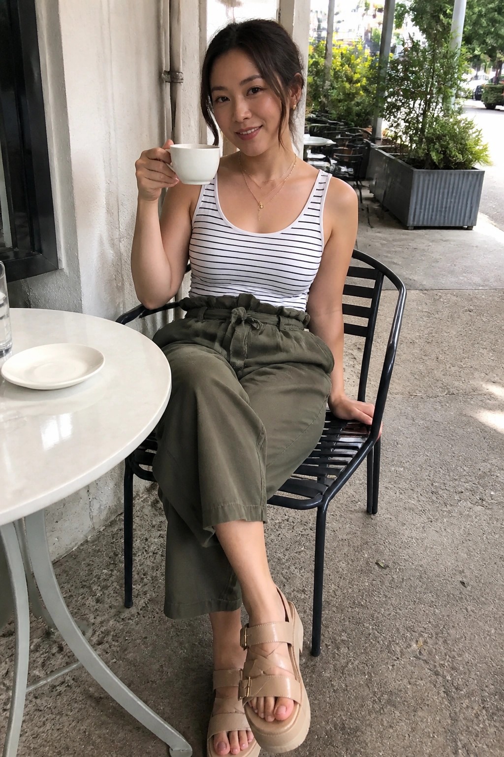 Woman in striped tank top and olive pants holding coffee cup at outdoor cafe