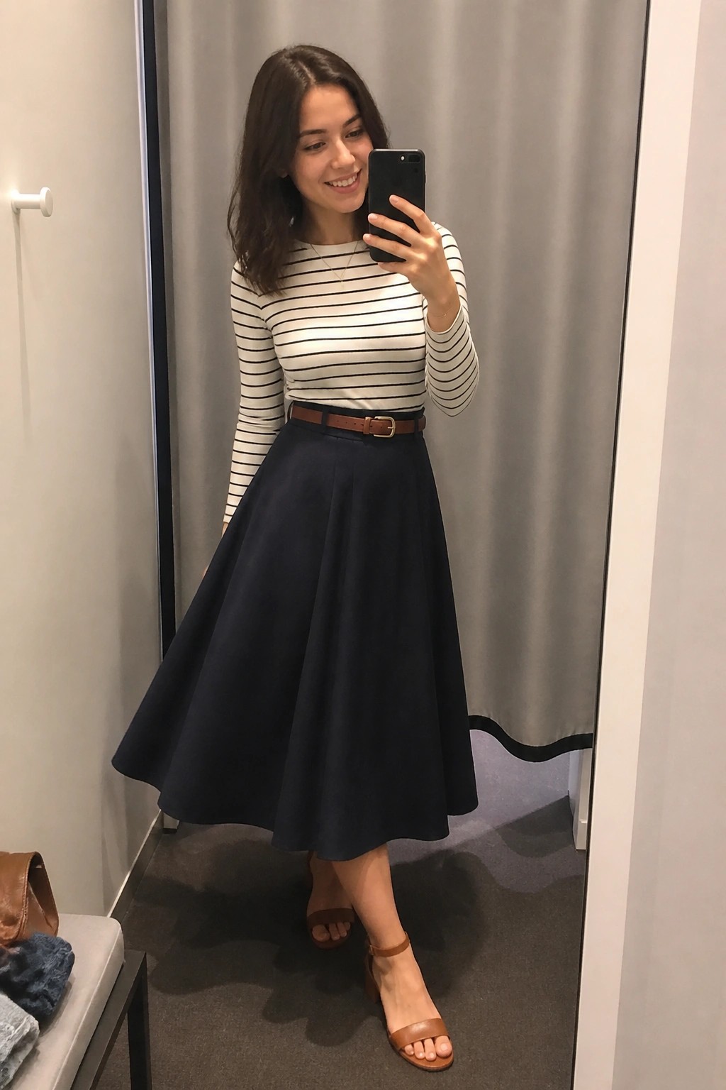 Woman in a fitted striped long sleeve top tucked into a navy A line midi skirt with brown belt and matching brown sandals taking a mirror selfie in a fitting room.