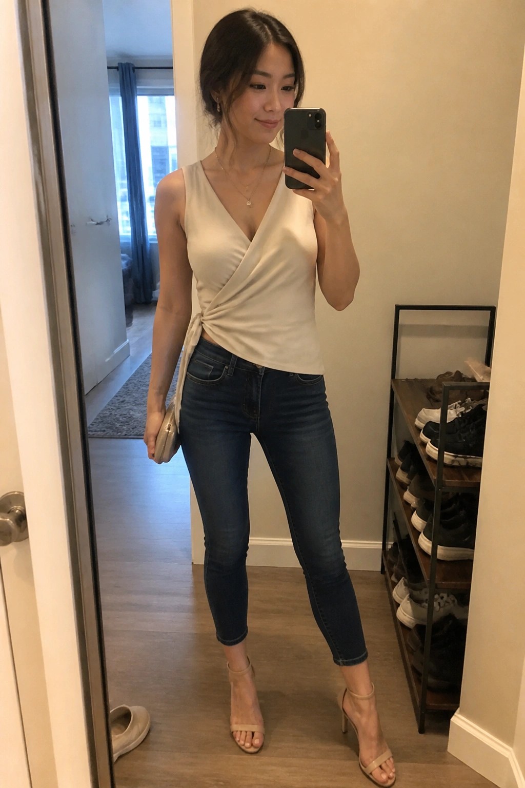 Woman in cream wrap top and jeans taking mirror selfie with phone.