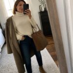 Woman wearing cream turtleneck sweater with dark jeans, beige coat draped over shoulders, beige ankle boots, and brown leather tote bag while holding a phone.
