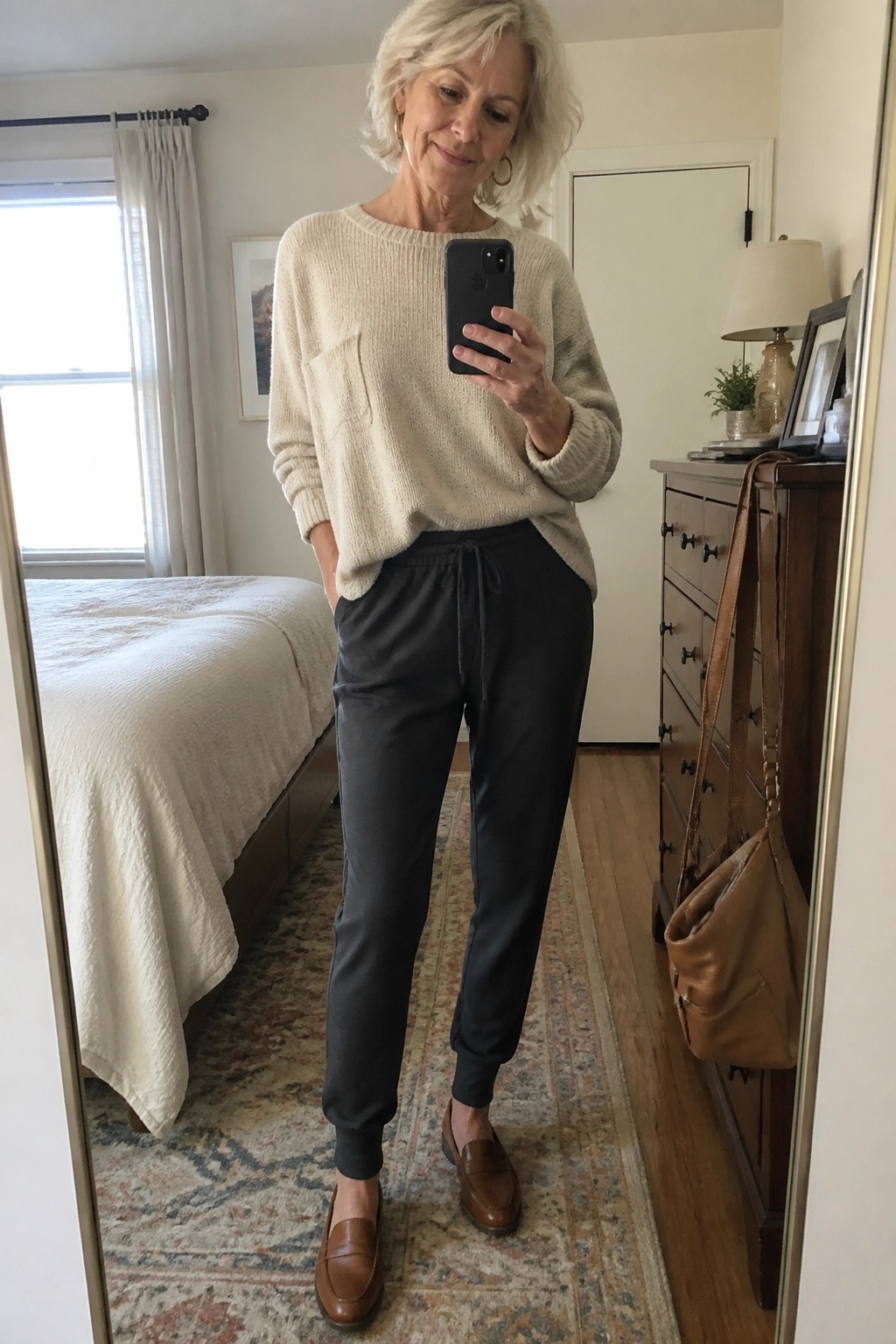 Short-haired woman in cream sweater and black joggers taking bedroom mirror selfie