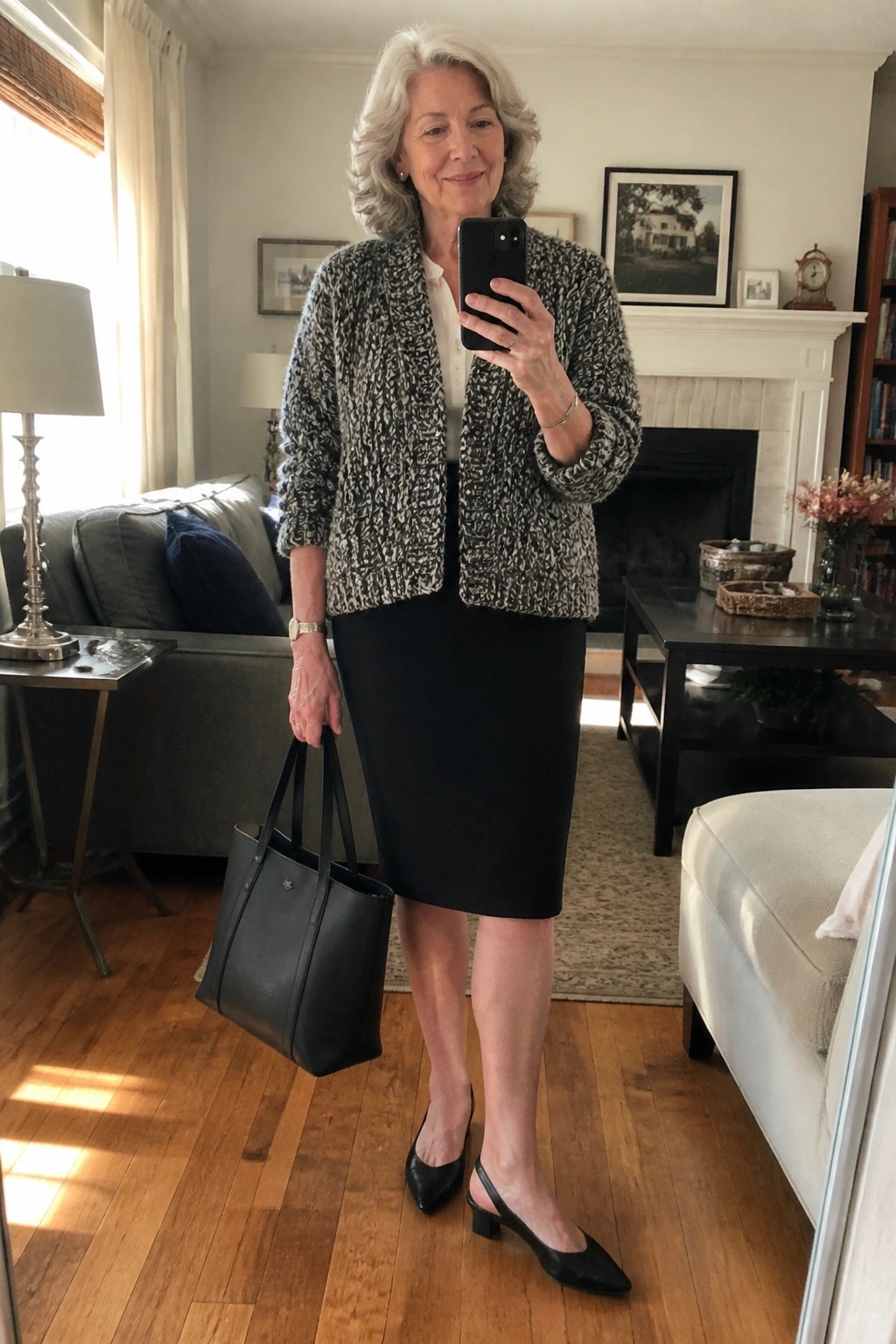 Gray-haired woman in marled jacket and black skirt holding phone and handbag in living room