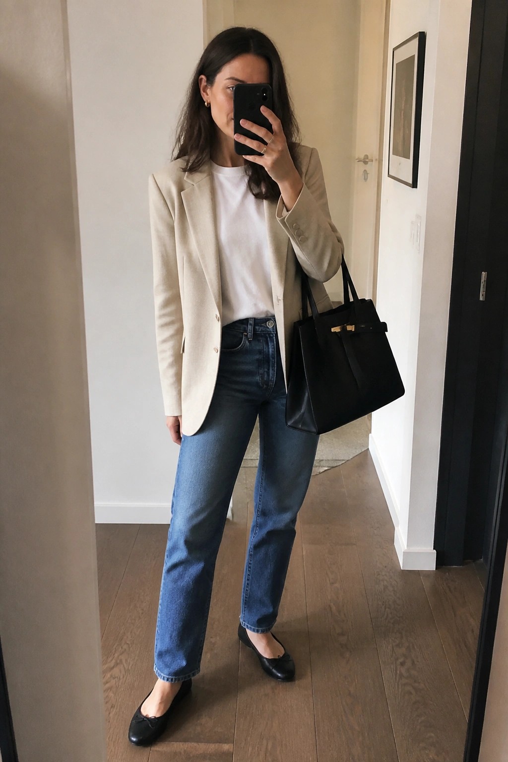 Woman in beige blazer and blue jeans taking mirror selfie with black handbag