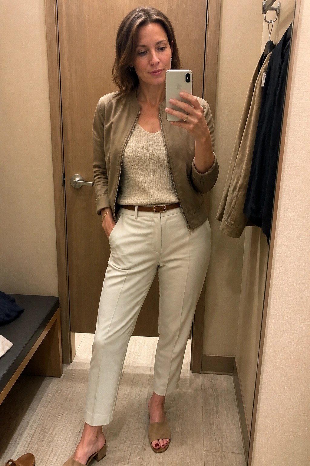 Woman taking a mirror selfie in a fitting room wearing a taupe bomber jacket zipped over a cream knit tank top, paired with light beige straight leg trousers, a brown leather belt, and beige open toe slides.