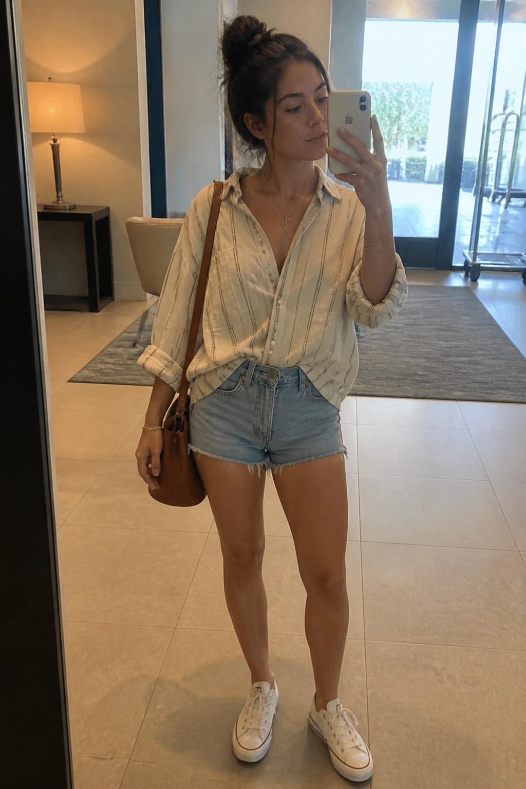 Woman with bun in striped shirt and denim shorts taking mirror selfie with phone