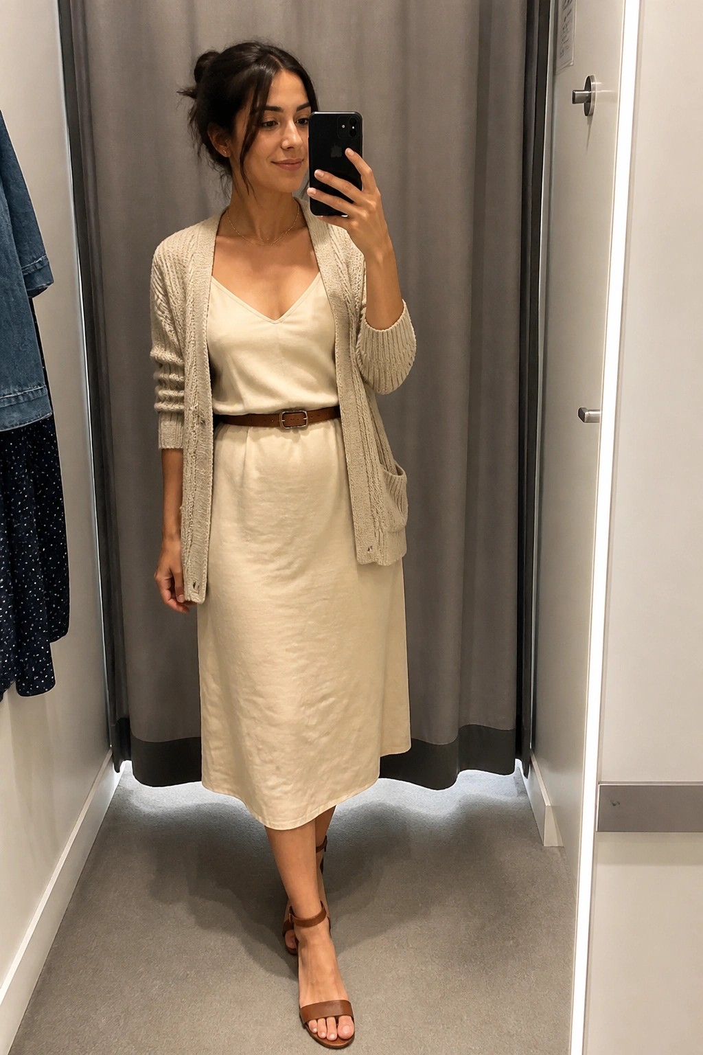 Woman in beige cardigan and cream dress taking mirror selfie in fitting room