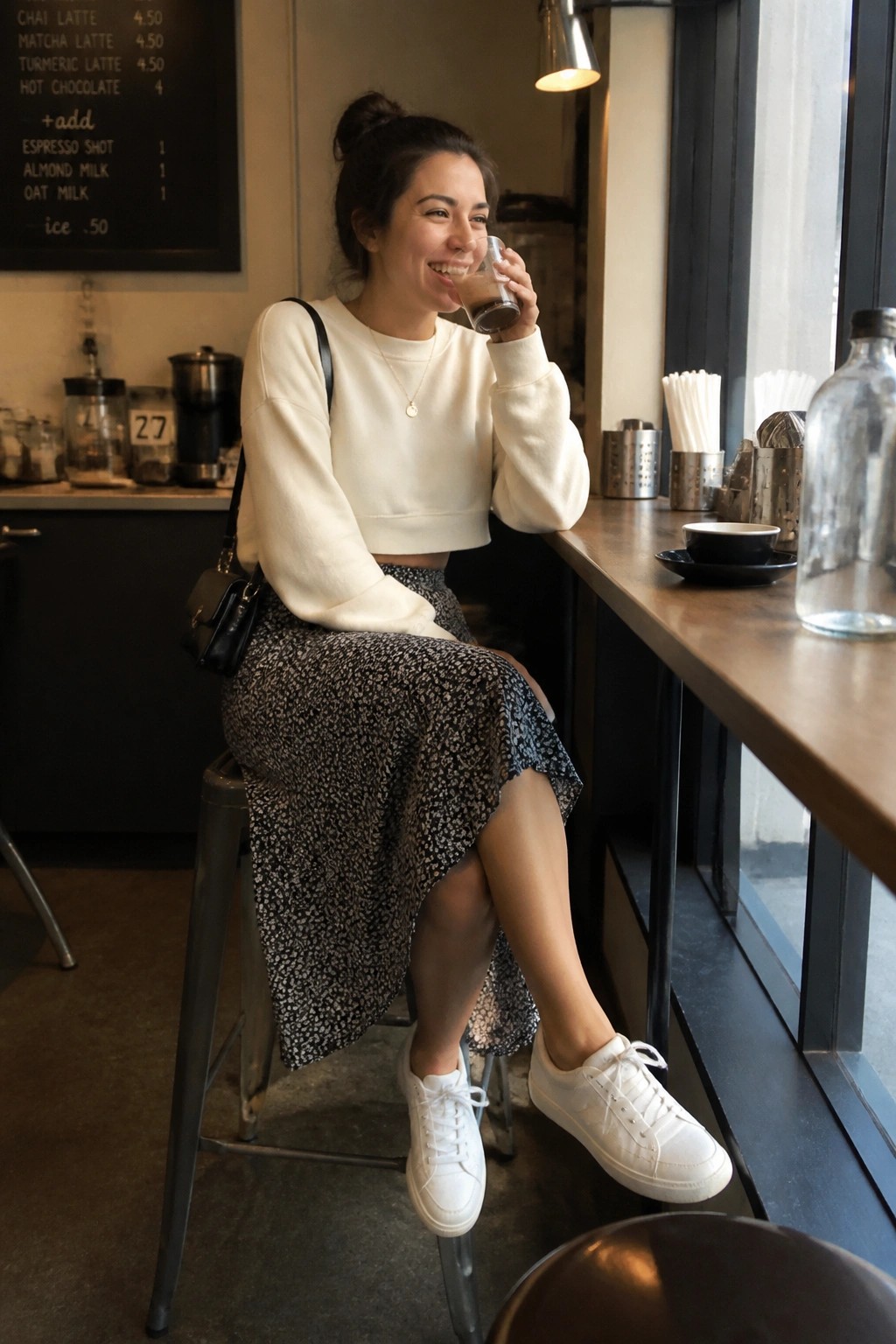 Woman in white crop top and floral skirt smiles while sipping iced coffee in cafe