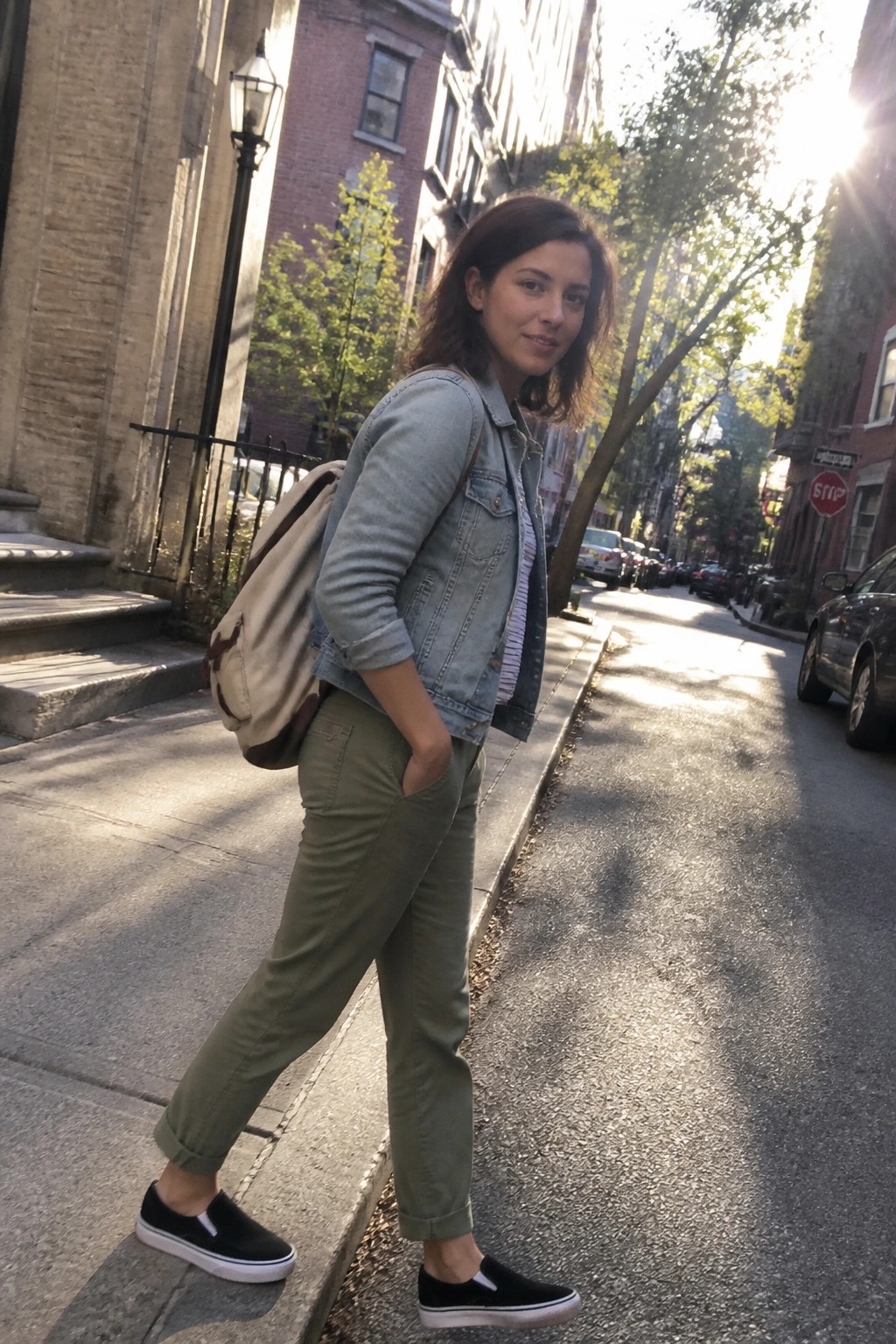 Woman in light denim jacket, olive pants, and backpack on sunny city sidewalk