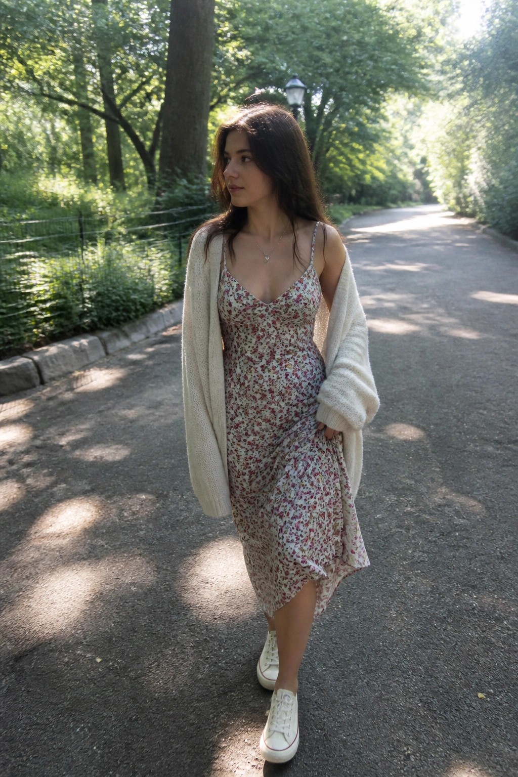 Woman wearing a cream open knit cardigan over a floral print slip dress with white lace-up sneakers on a paved path.