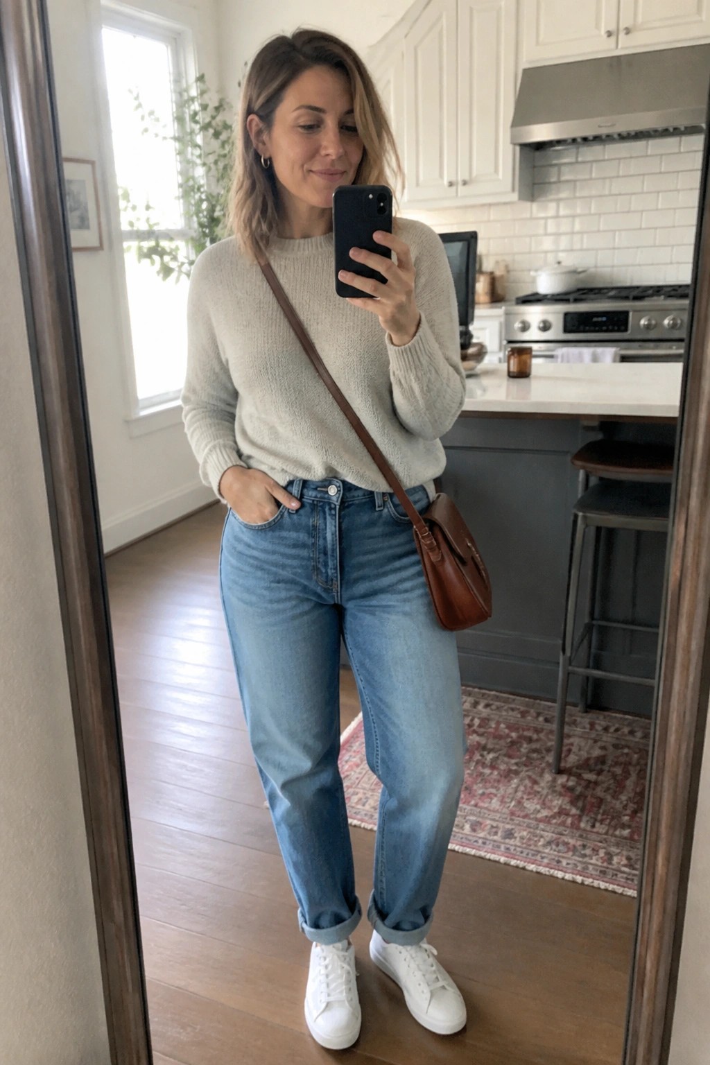 Woman in mirror selfie wearing light beige fuzzy knit sweater, blue cuffed jeans, white sneakers, and brown leather crossbody bag.