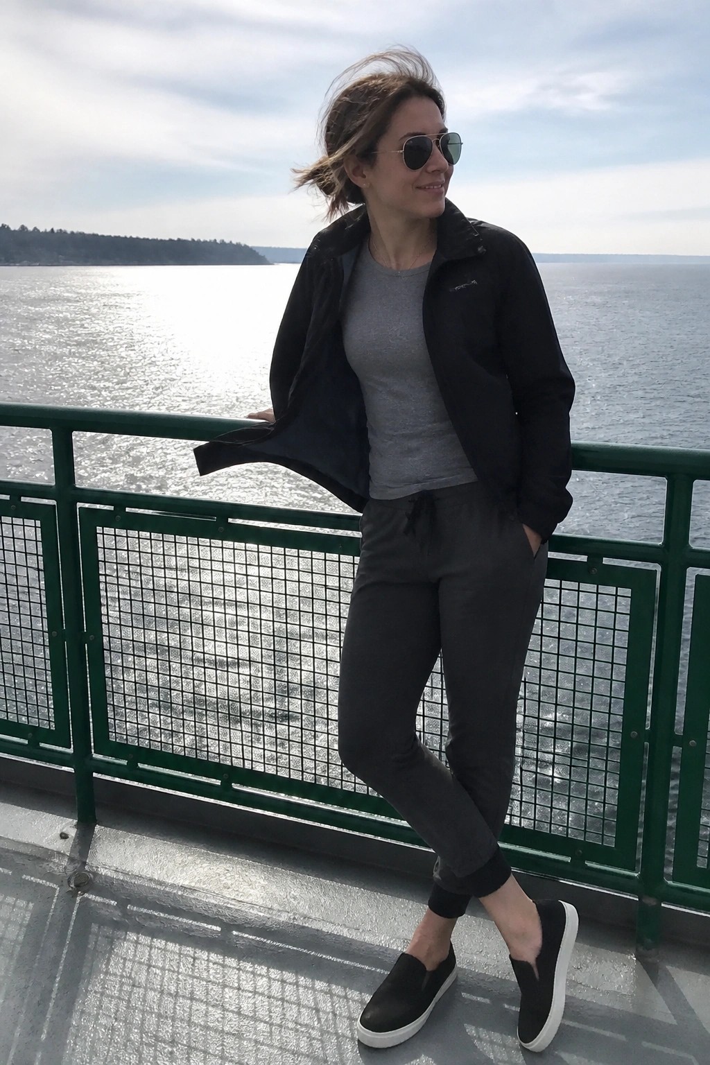 Woman wearing an open black zip jacket over a gray t-shirt paired with dark gray jogger pants and black slip on sneakers with white soles.