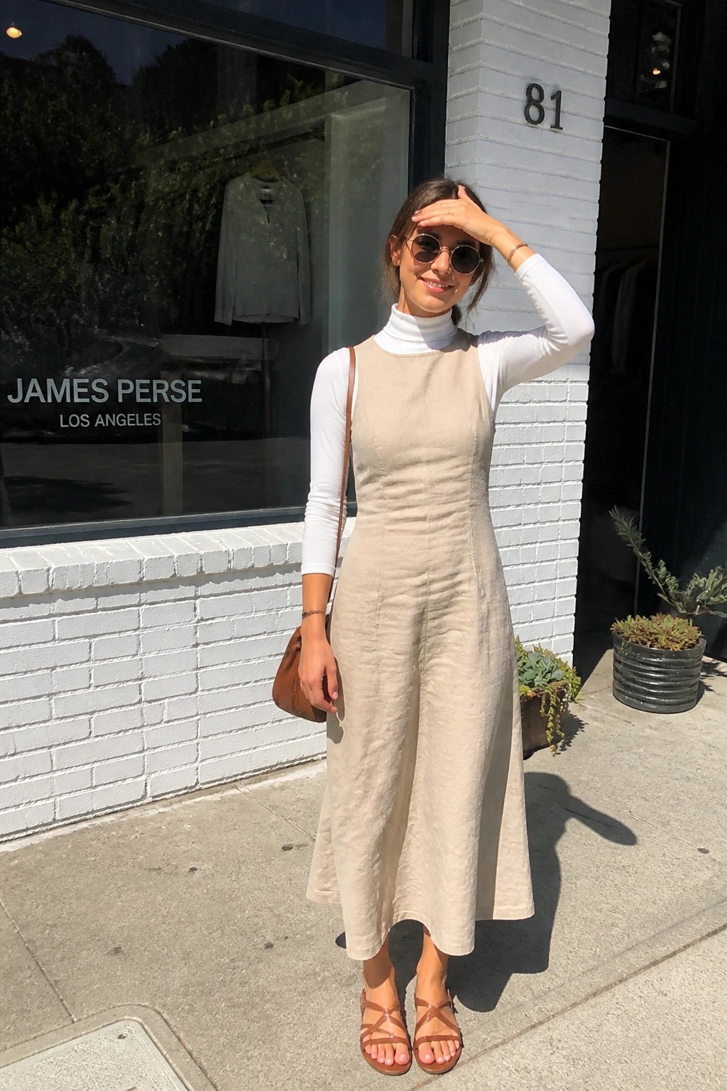 Woman in beige linen dress over white turtleneck outside James Perse storefront on sunny sidewalk.