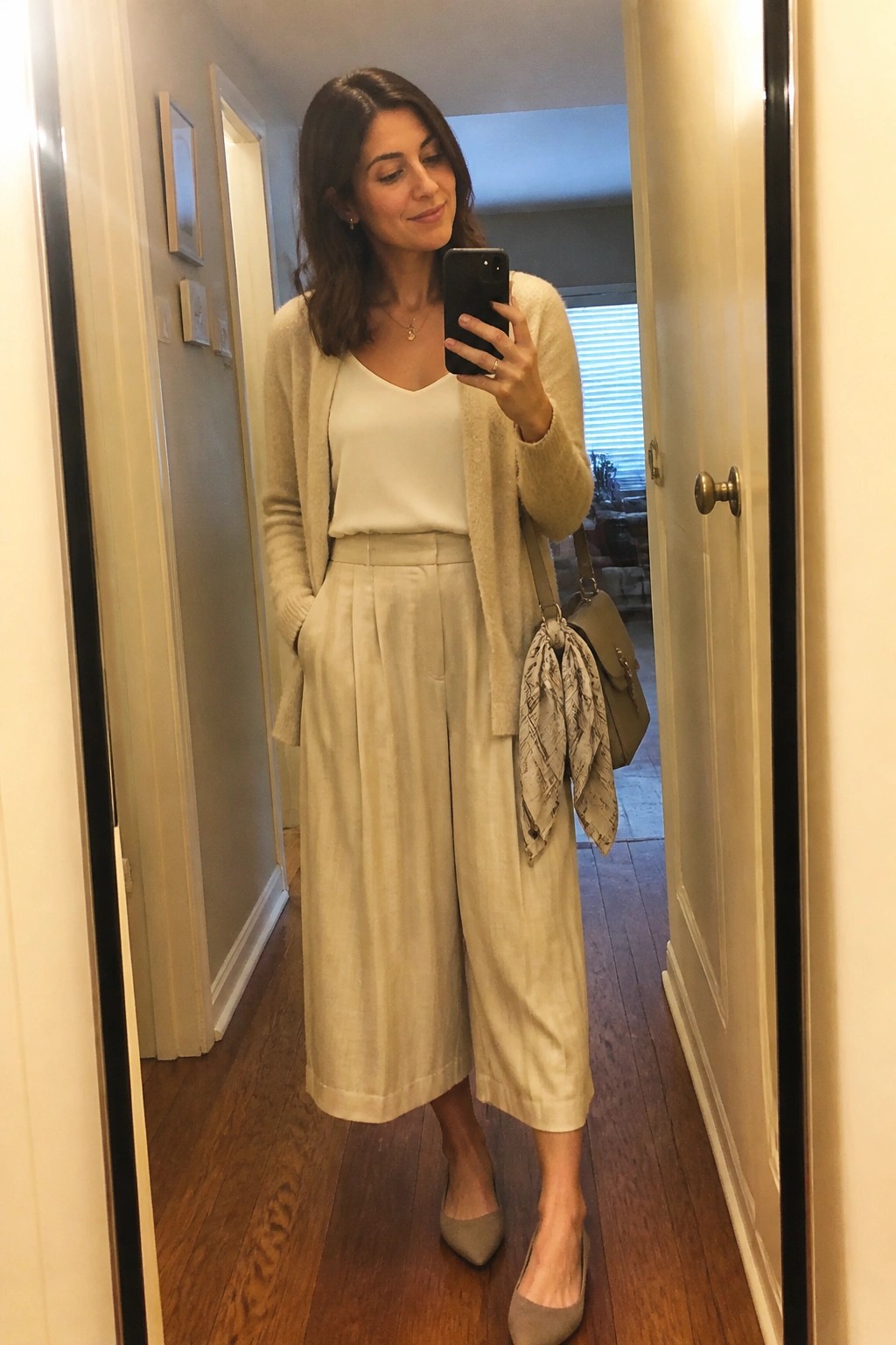 Woman in beige cardigan and wide-leg pants taking hallway mirror selfie with handbag