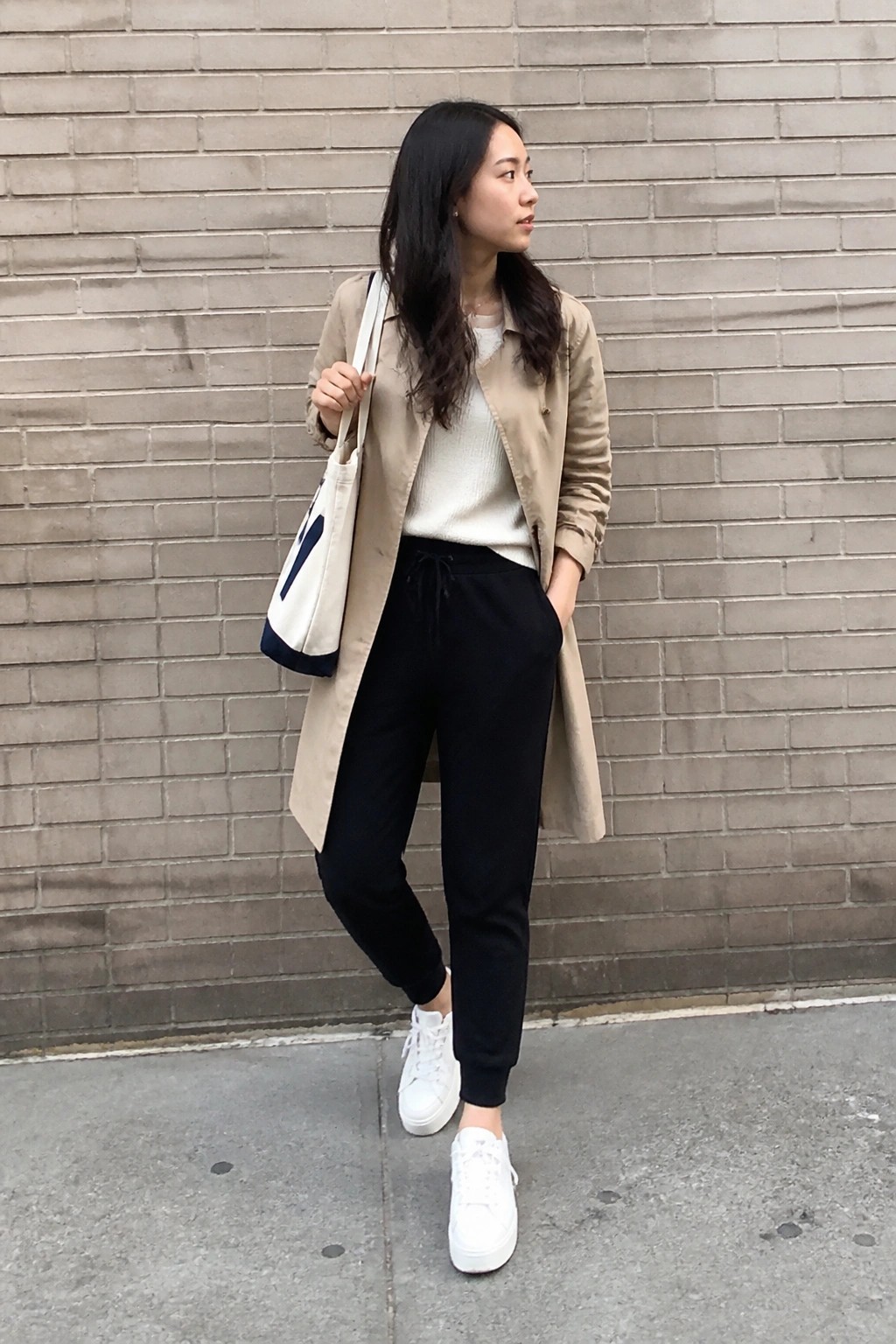 Woman in beige trench coat, white sweater, black joggers holds tote bag by brick wall.