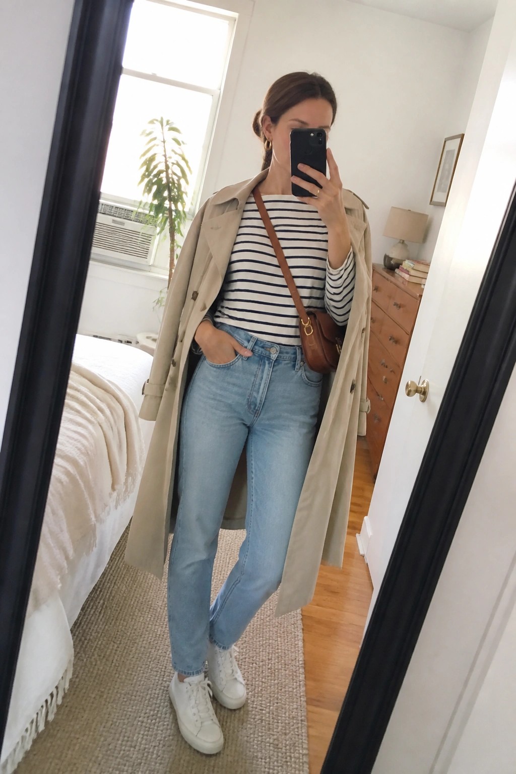Woman in beige trench coat and striped shirt taking bedroom mirror selfie