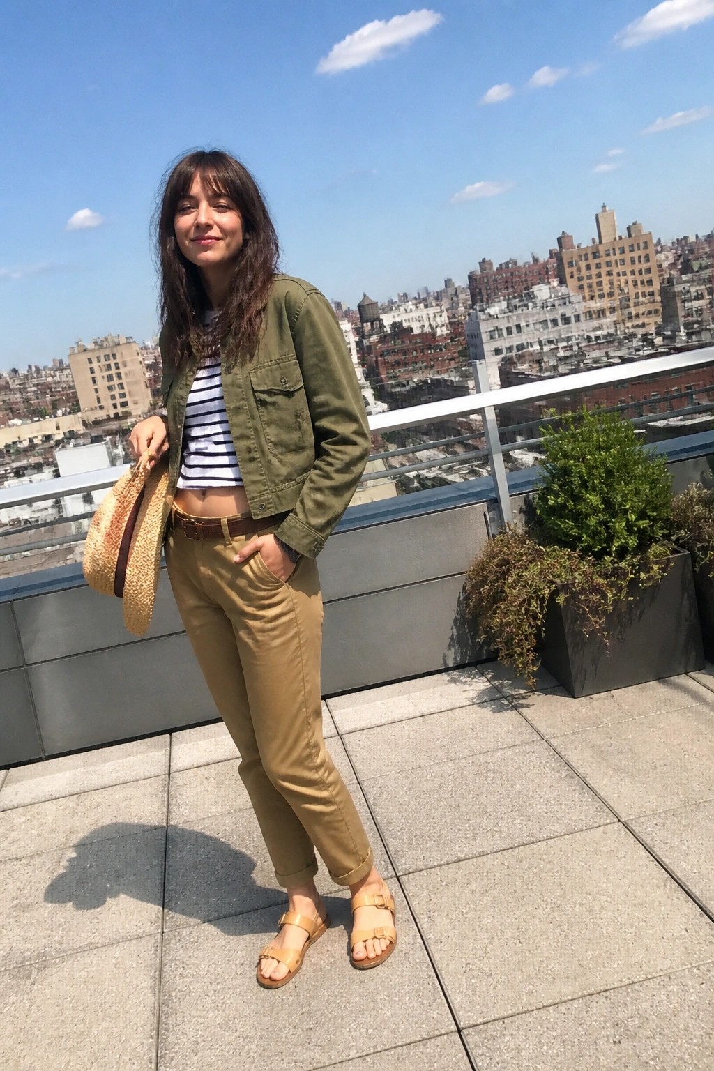 Woman in green jacket and khaki pants on sunny city rooftop holding straw hat.
