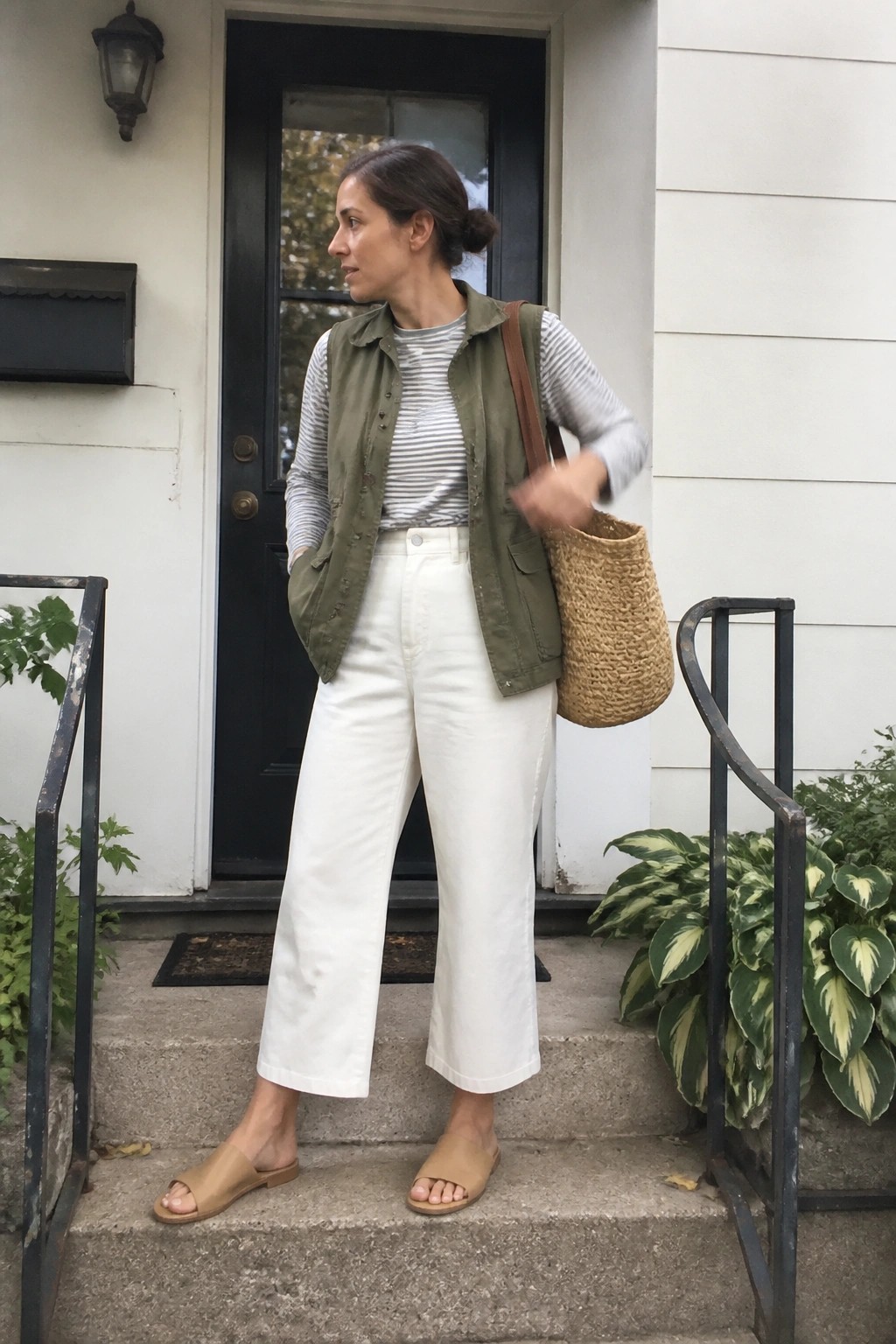 Woman in green vest, striped shirt, white pants holding woven bag on house steps