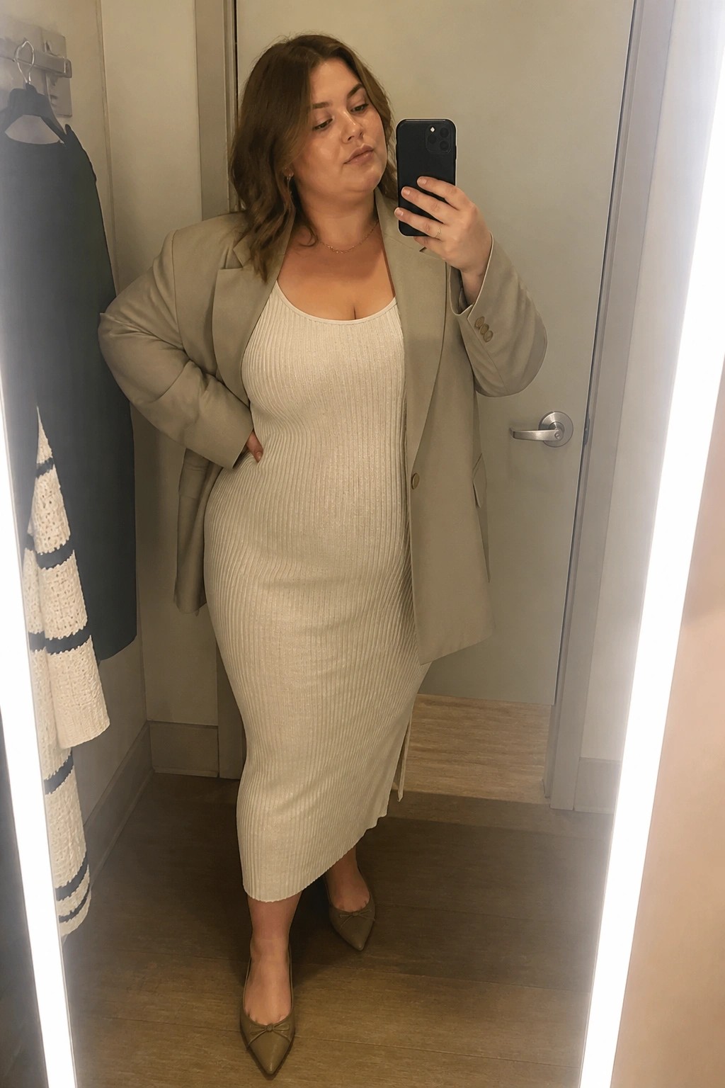 Woman in beige blazer and ribbed dress taking mirror selfie in fitting room