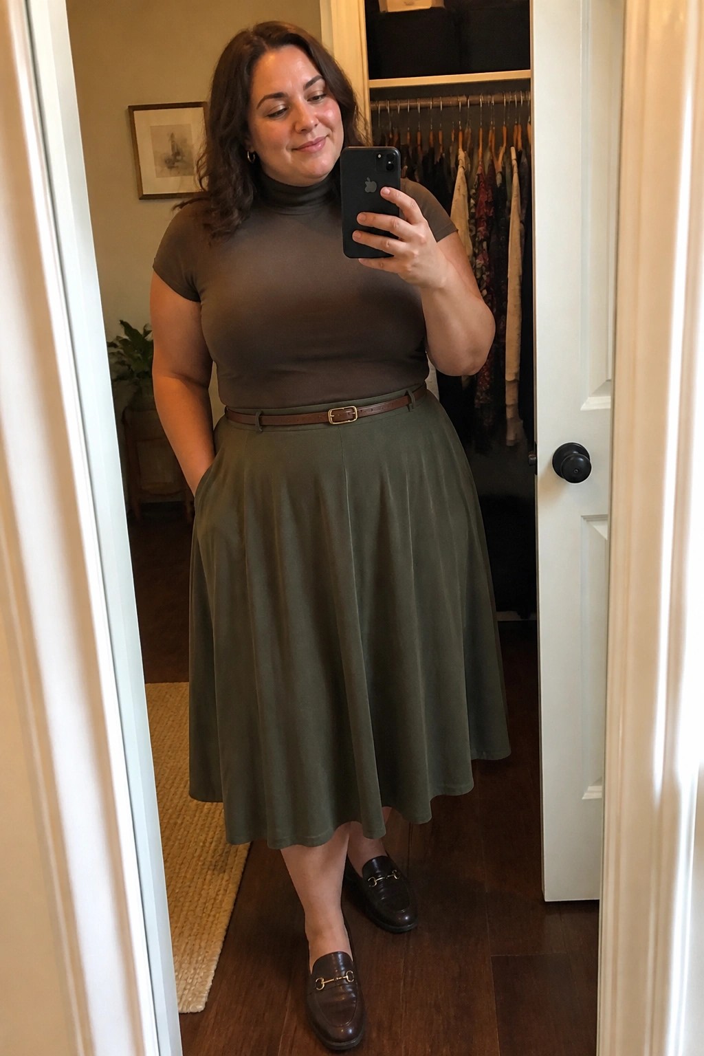 Woman in brown turtleneck and olive skirt taking mirror selfie with phone.