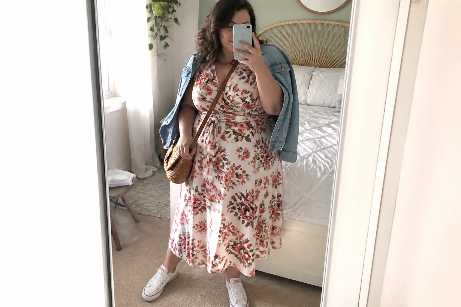 Woman in yellow floral wrap dress, denim jacket, and white Converse taking bedroom mirror selfie