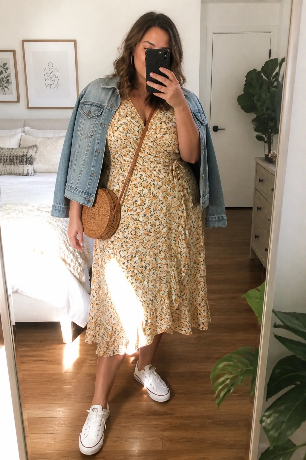 Woman in yellow floral wrap dress, denim jacket, and white Converse taking bedroom mirror selfie