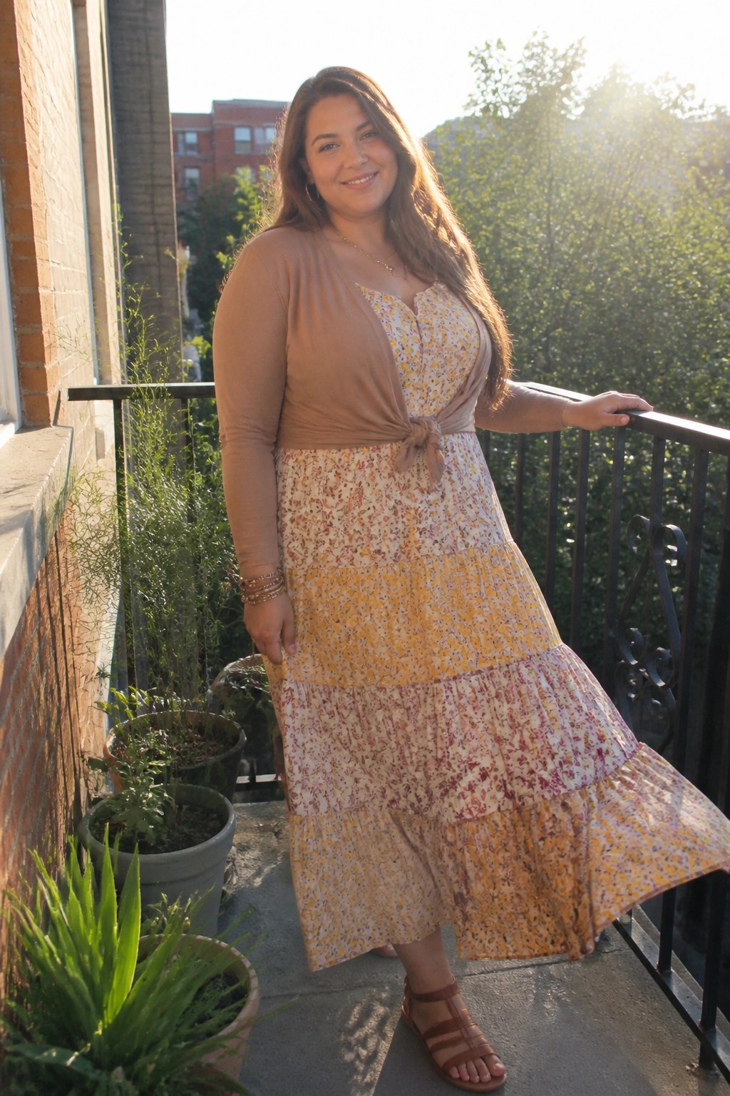 Plus size woman in a yellow pink floral tiered midi dress layered with a tied tan long sleeve top and brown strappy sandals on a balcony.