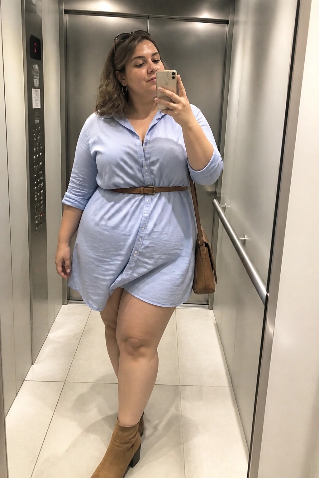 Woman in light blue shirt dress taking selfie in elevator with brown belt and boots