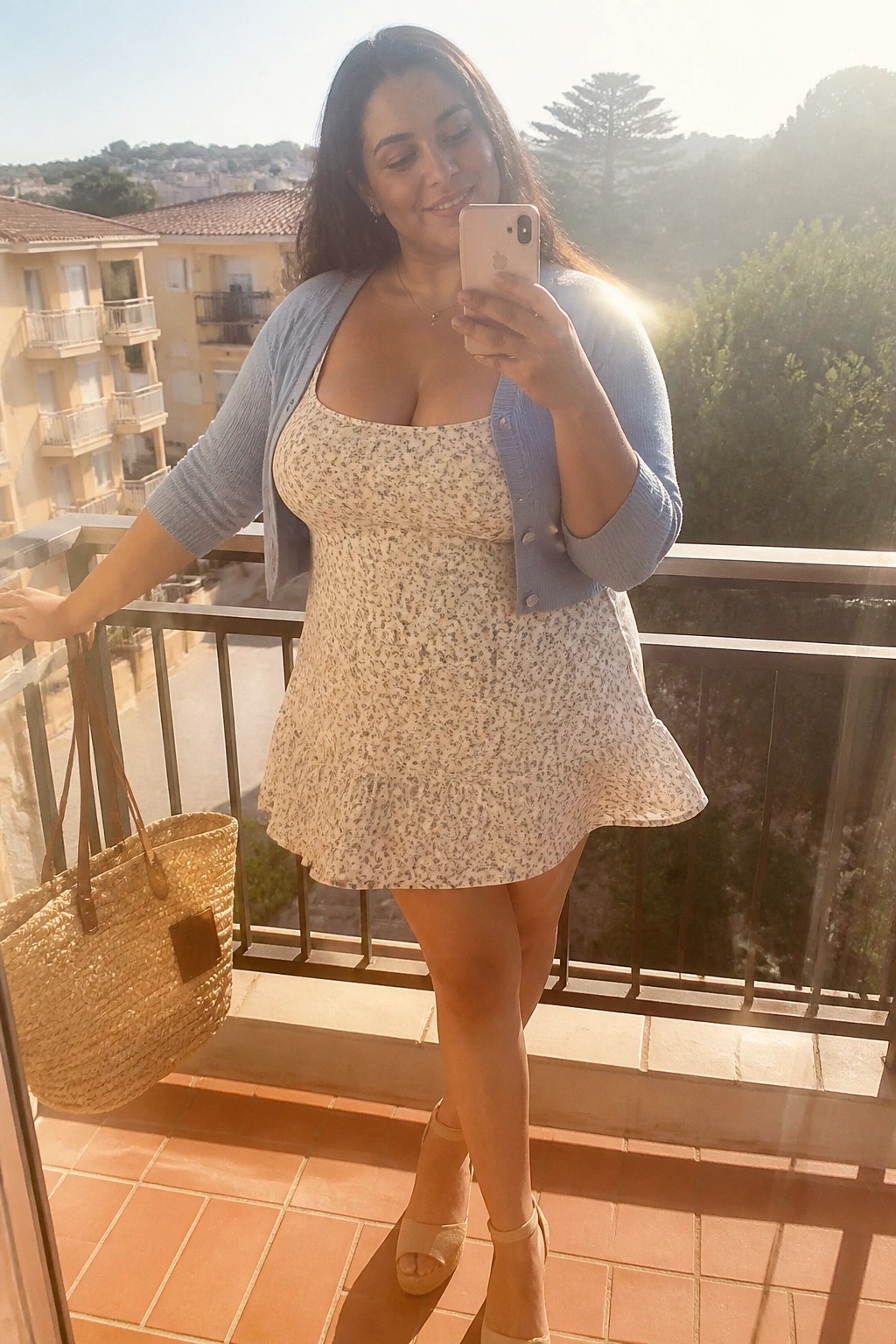 Woman in floral dress and blue cardigan taking selfie on sunny balcony with straw bag